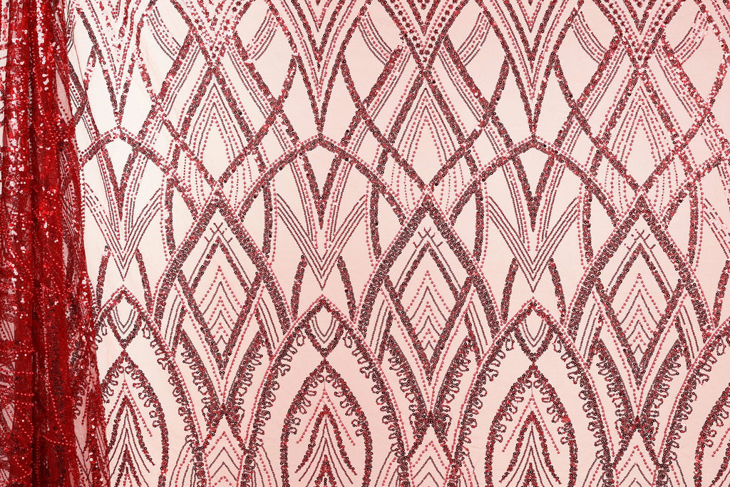a close-up view of a fabric with a repeating pattern of red sequin designs. The pattern consists of various shapes and lines, creating a visually striking and intricate design. The fabric appears to be a pink or peach-colored material, which contrasts with the red sequin pattern. The image does not contain any text or other discernible objects, and the focus is solely on the fabric and its design.