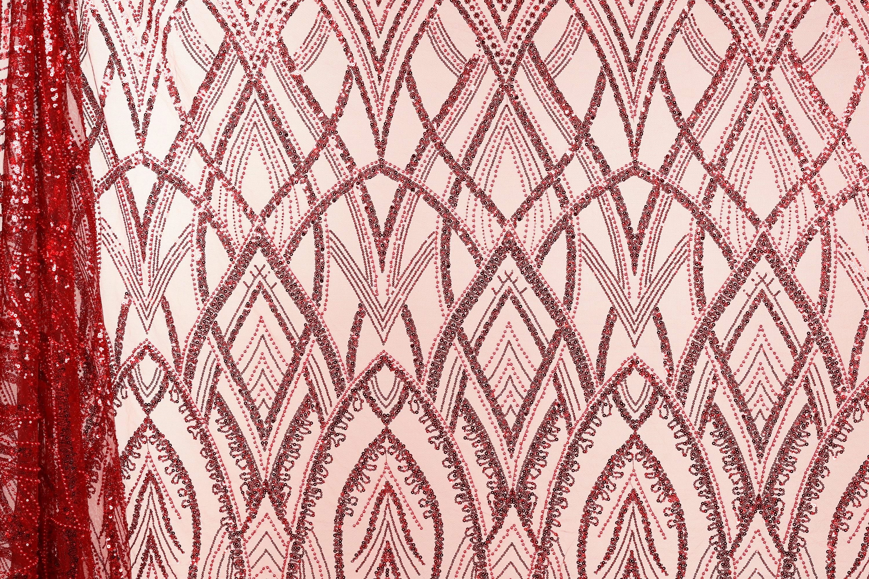 a close-up view of a fabric with a repeating pattern of red sequin designs. The pattern consists of various shapes and lines, creating a visually striking and intricate design. The fabric appears to be a pink or peach-colored material, which contrasts with the red sequin pattern. The image does not contain any text or other discernible objects, and the focus is solely on the fabric and its design.