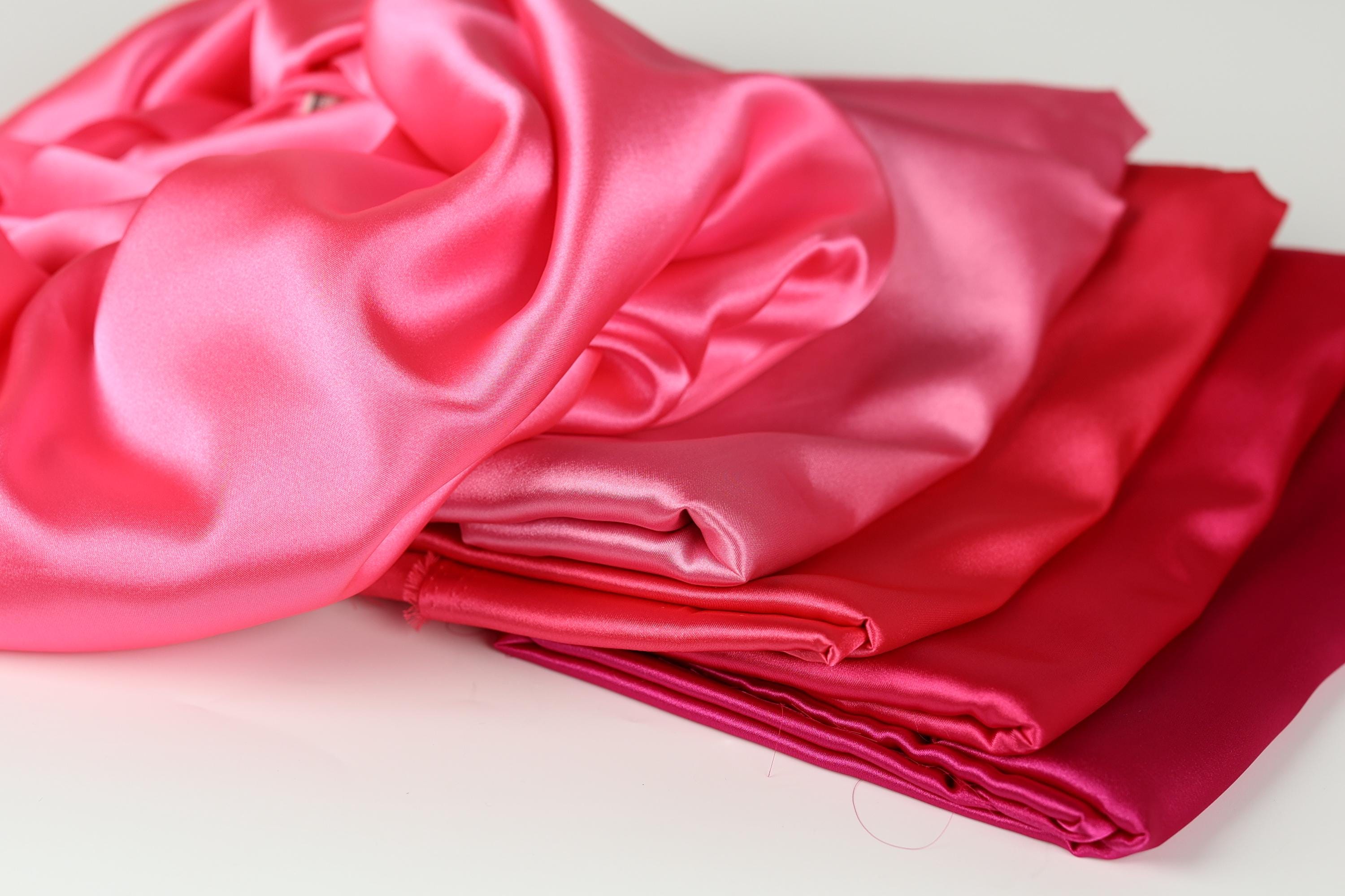 A pile of soft, shiny pink satin fabric is displayed on a white surface.
