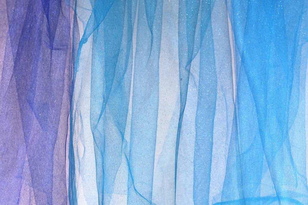 a close-up view of a piece of blue and purple fabric, with the fabric appearing to be made of sheer material. The fabric has a textured and layered appearance, with vertical lines and stripes creating a sense of depth and movement. The colors of the fabric are predominantly blue and purple, with hints of white and gray adding contrast and balance to the overall composition.