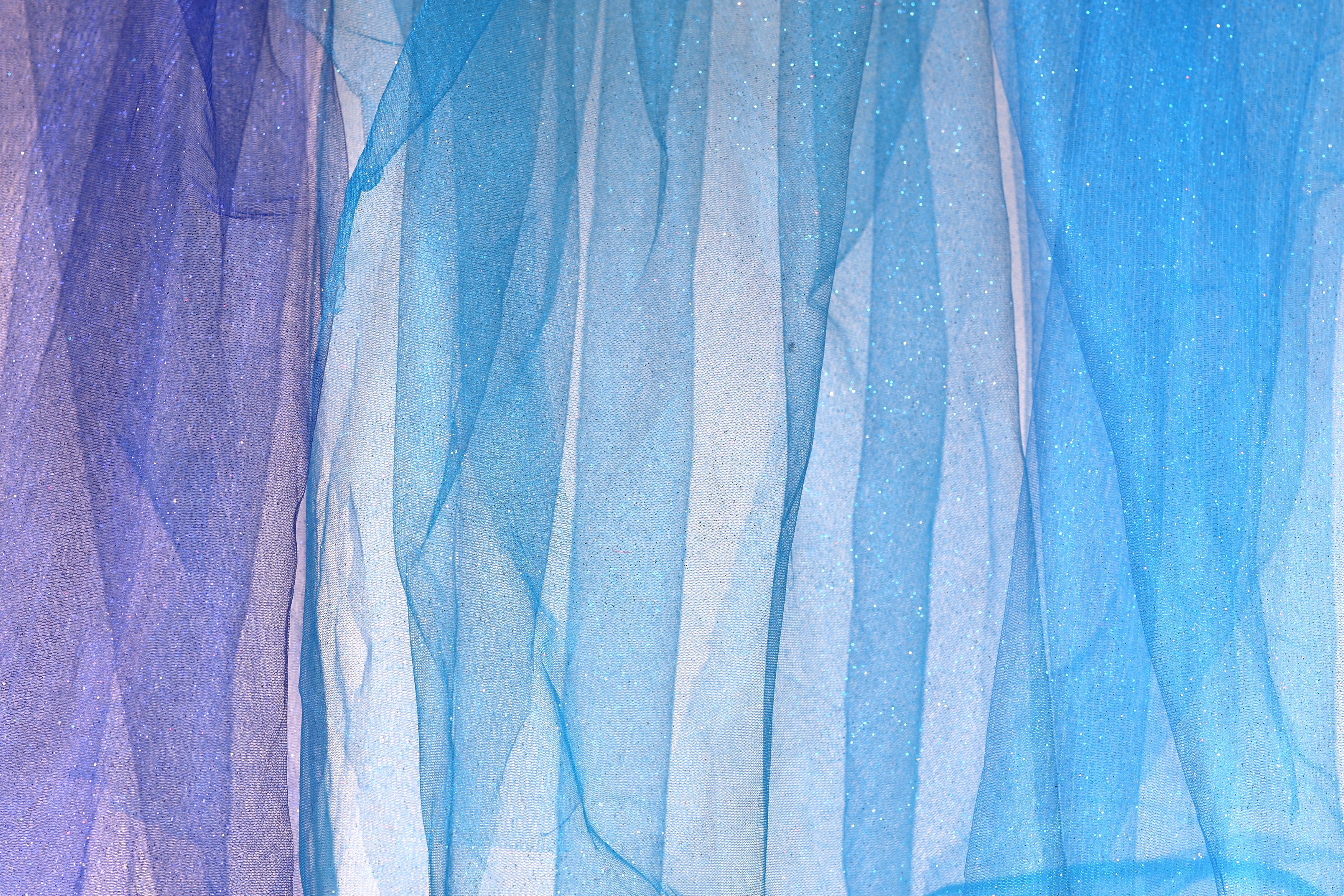 a close-up view of a piece of blue and purple fabric, with the fabric appearing to be made of sheer material. The fabric has a textured and layered appearance, with vertical lines and stripes creating a sense of depth and movement. The colors of the fabric are predominantly blue and purple, with hints of white and gray adding contrast and balance to the overall composition.