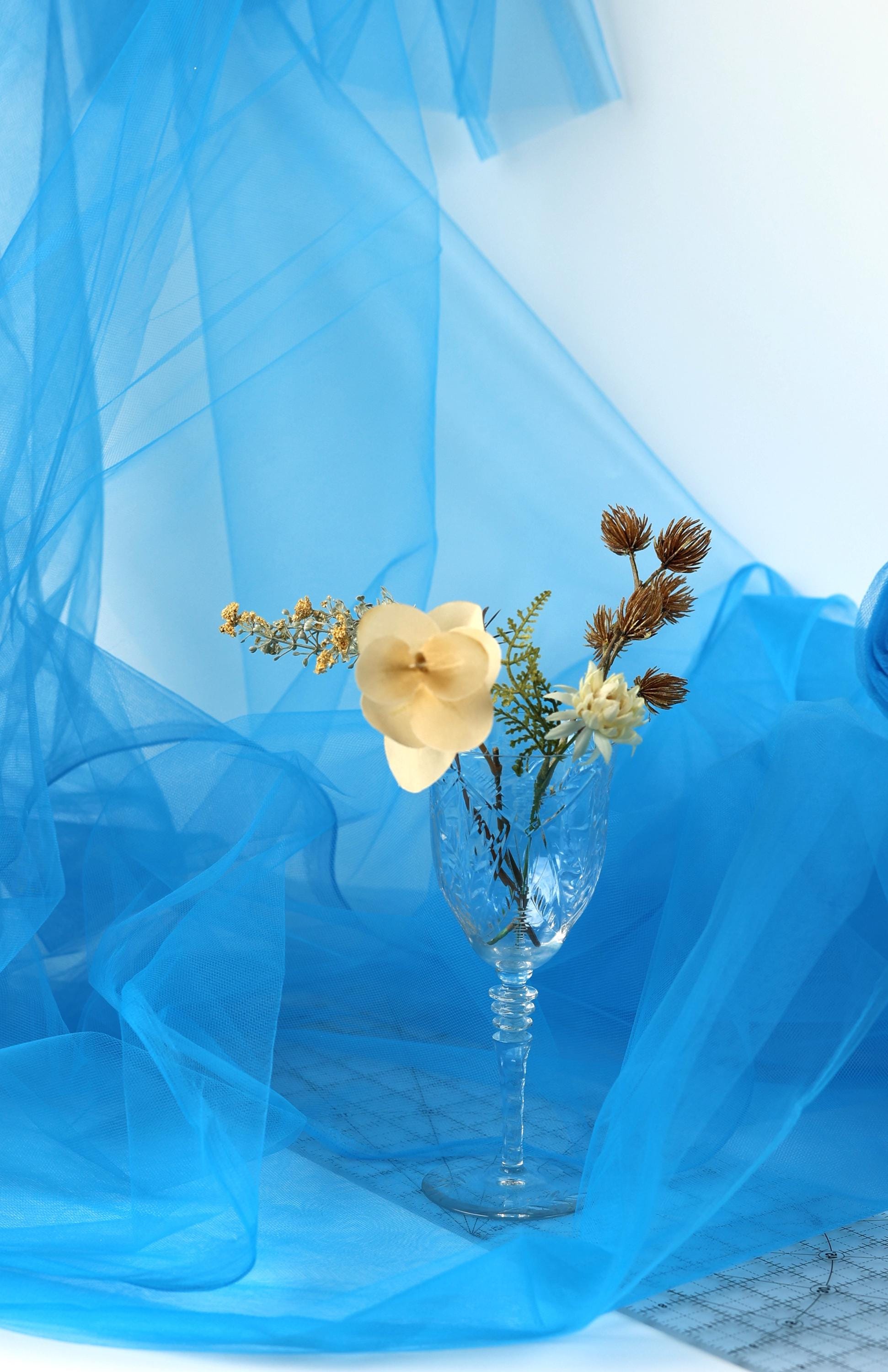 A glass vase filled with a variety of flowers sits on a blue fabric background.