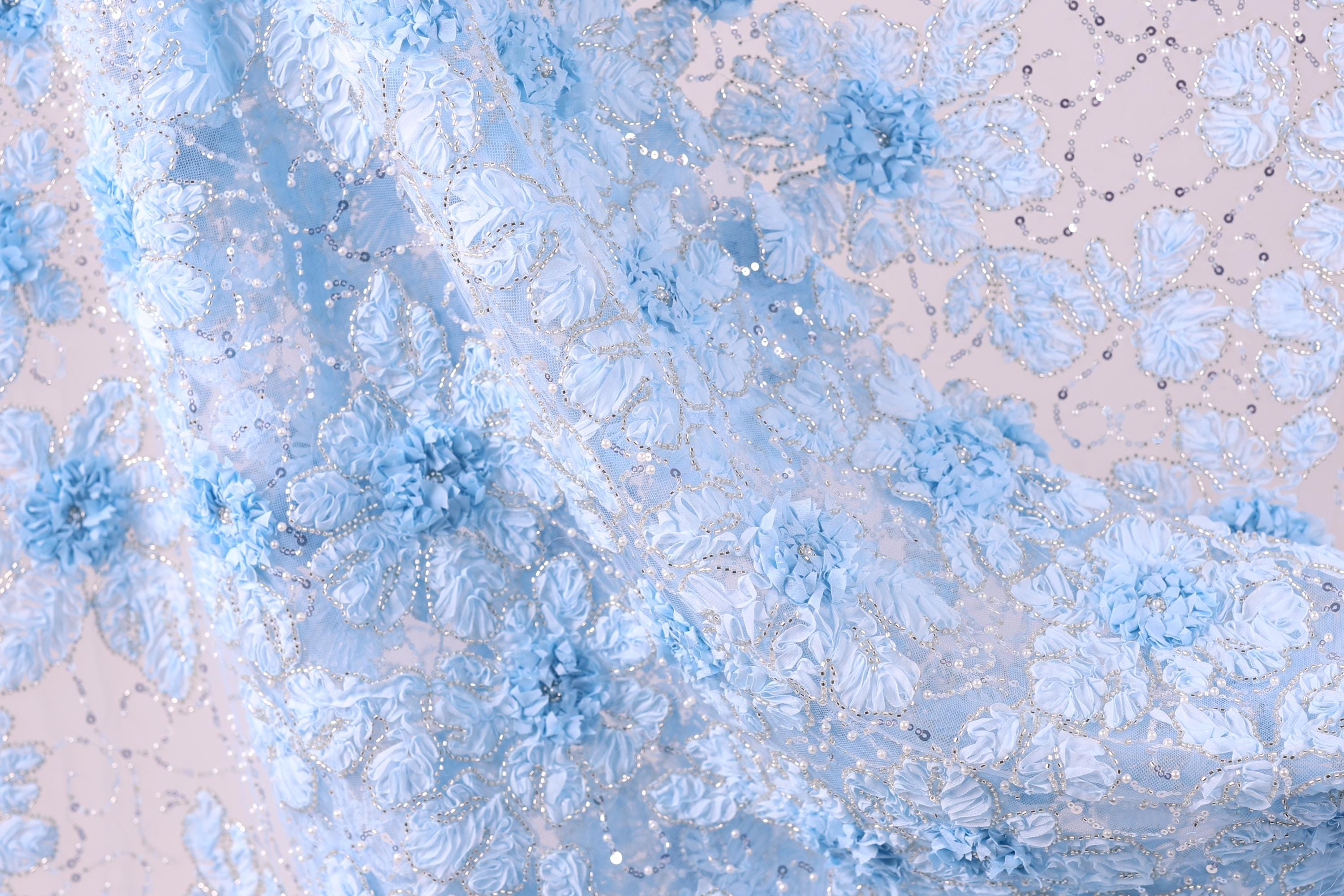 The image is a close-up of a delicate, intricately designed fabric with a floral pattern. The fabric appears to be made of a light blue material, possibly lace or chiffon, with small white flowers or leaves scattered throughout the design. The fabric has a soft, flowing texture, and the intricate details of the floral pattern create a visually appealing and delicate appearance.