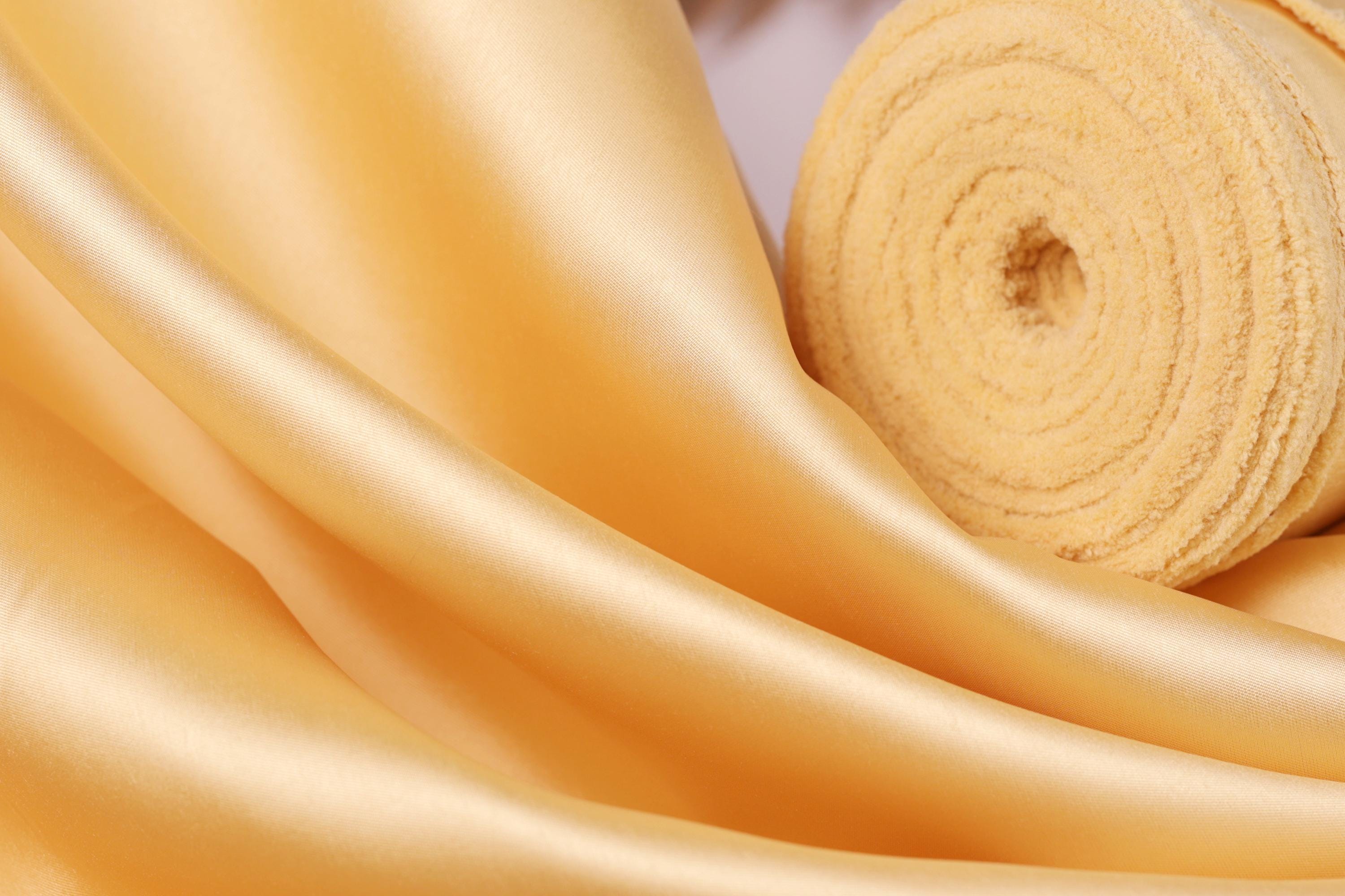 a close-up view of a soft, shiny fabric with a golden hue. The fabric appears to be made of silk or a similar luxurious material, and it is folded or rolled up, creating a visually appealing texture.