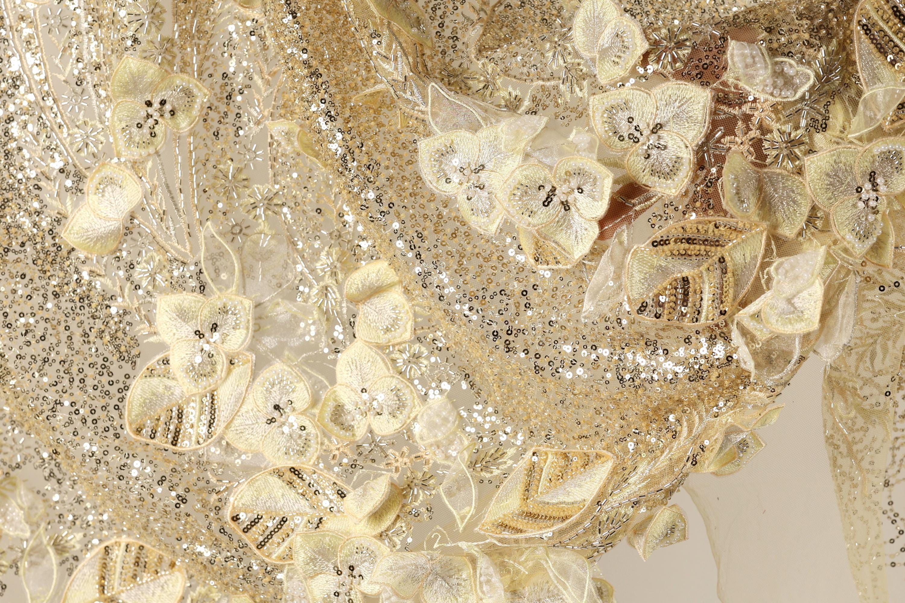 The image is a close-up of a delicate, shimmering fabric with a floral pattern. The fabric is adorned with numerous small, intricately designed flowers and sequins, creating a visually striking and elegant appearance.