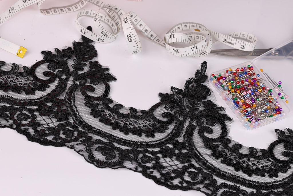 a close-up view of a black lace trim with intricate floral patterns, along with a pair of scissors, a measuring tape, and a container of colorful pins.