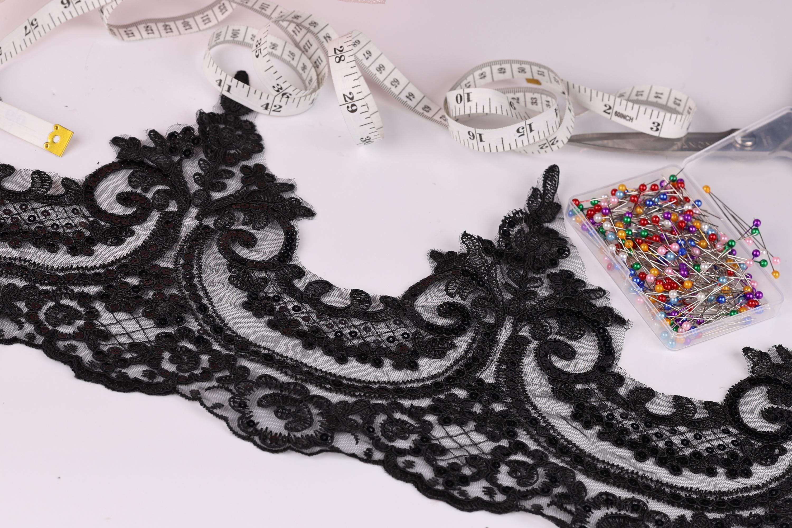 a close-up view of a black lace trim with intricate floral patterns, along with a pair of scissors, a measuring tape, and a container of colorful pins.