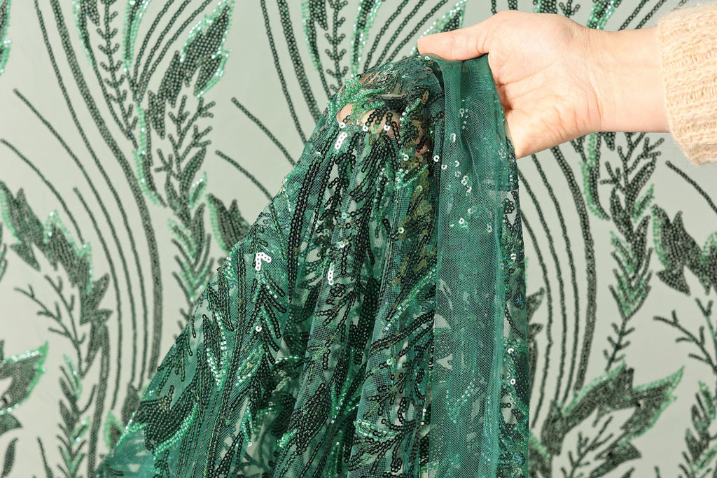 A hand is holding a piece of green sequined fabric with a pattern of green leaves and stems.