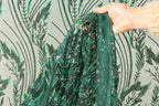 A hand is holding a piece of green sequined fabric with a pattern of green leaves and stems.