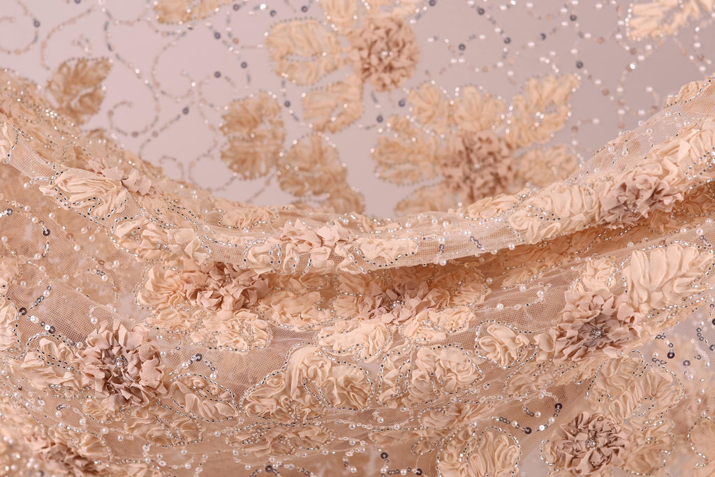 a close-up view of a delicate, beige-colored fabric with a floral pattern. The fabric appears to be made of lace or a similar delicate material, with small, intricately designed flowers or leaves adorning the surface. The overall appearance of the fabric is soft and delicate, with a sense of elegance and sophistication.
