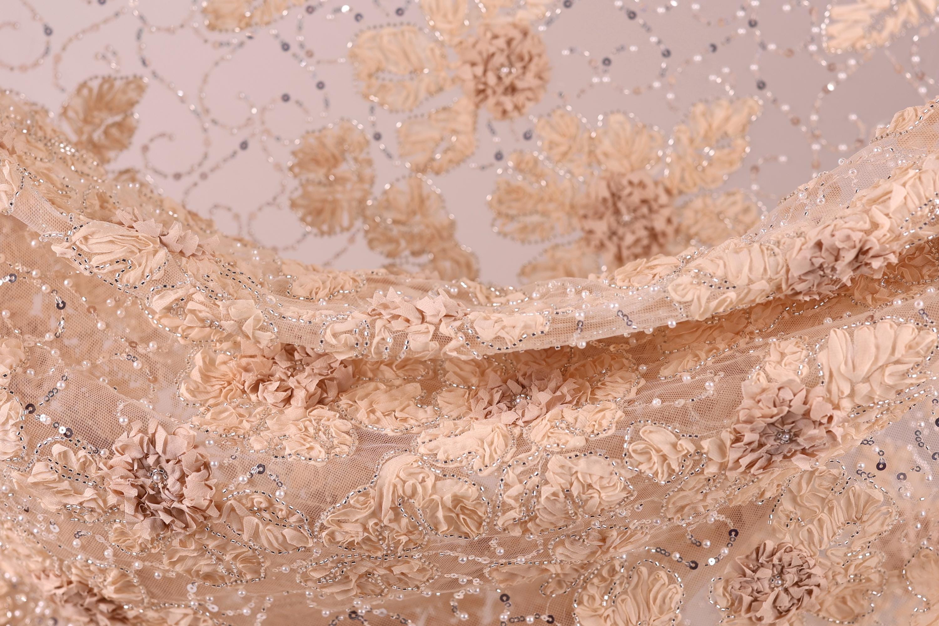 a close-up view of a delicate, beige-colored fabric with a floral pattern. The fabric appears to be made of lace or a similar delicate material, with small, intricately designed flowers or leaves adorning the surface. The overall appearance of the fabric is soft and delicate, with a sense of elegance and sophistication.