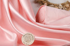 a pink satin fabric with a sewing artisan logo in the foreground, and a small rolled-up fabric piece in the background.