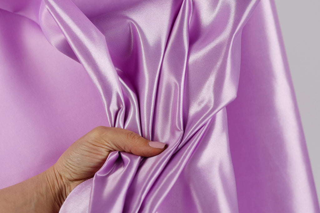 A hand is holding a piece of shiny, purple satin fabric, which appears to be a close-up view of the material.