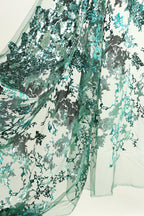 a close-up view of a fabric with a floral pattern in shades of green and blue. The fabric appears to be made of sheer material, with a textured and intricate design that resembles a lace or chiffon pattern. The colors and details of the fabric create a visually appealing and delicate appearance.