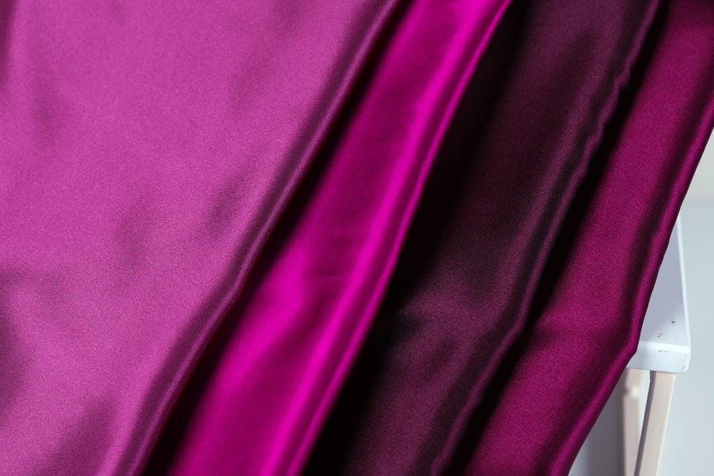 a close-up view of a fabric with three distinct shades of purple, ranging from a deep, rich purple to a lighter, more vibrant shade. The fabric appears to be made of a shiny, satin-like material, giving it a luxurious and elegant appearance.