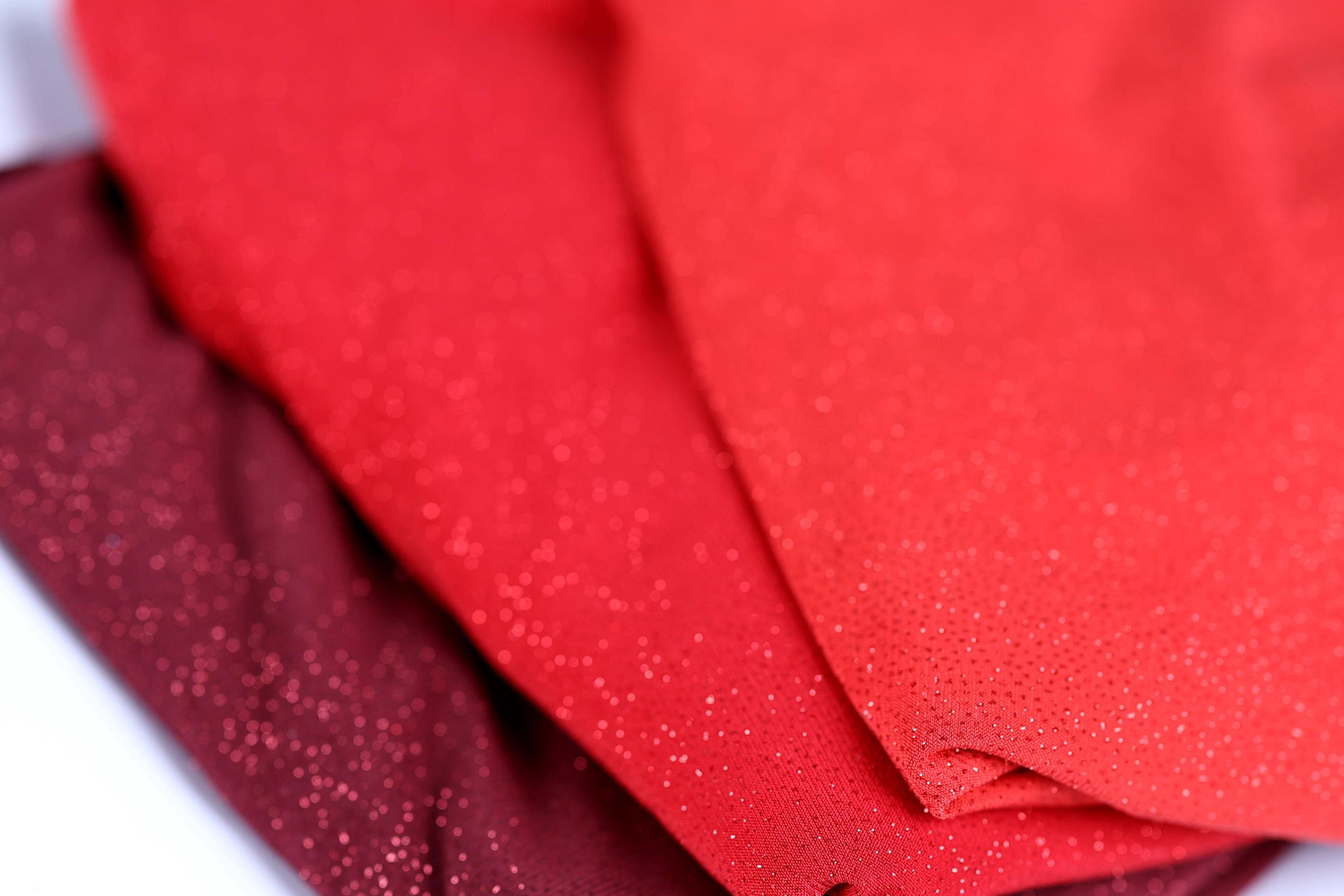 a close-up view of three fabric swatches, with the middle one being a vibrant red color and the other two being a darker red shade. The fabric appears to have a glittery or sparkly texture, adding a touch of sparkle to the overall appearance.