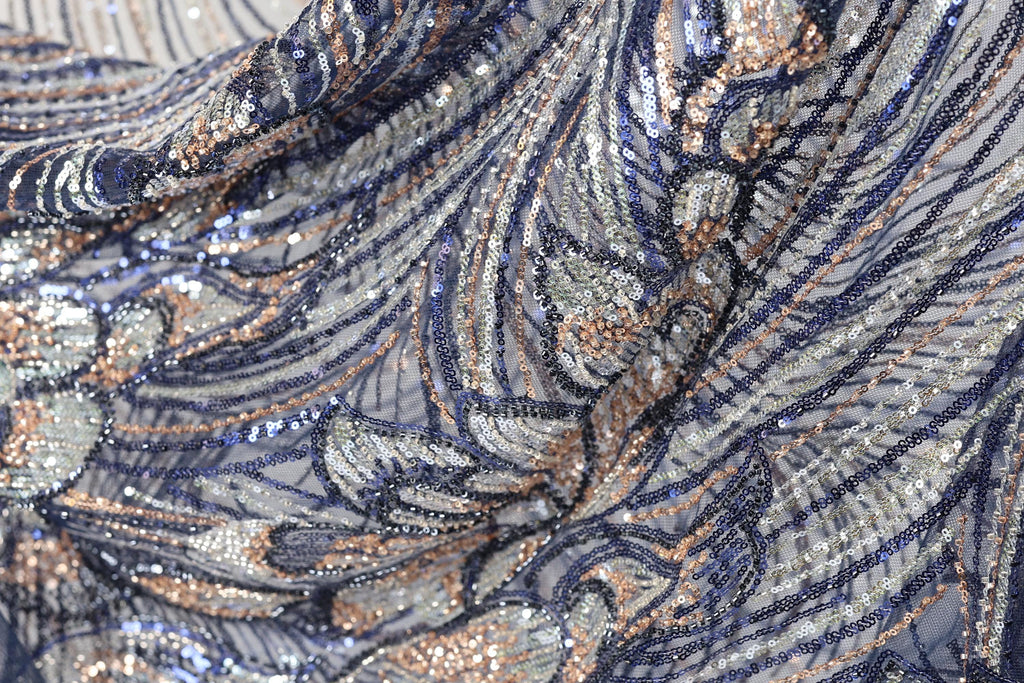 a close-up view of a fabric with a complex, intricate pattern of sequins and threads in shades of blue, silver, and gold. The fabric appears to be a piece of clothing or textile, with the intricate design and shimmering sequins creating a visually striking and detailed appearance.