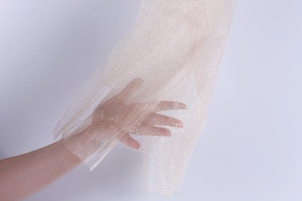 A hand is holding a piece of light-colored fabric, possibly a sheer or sheer-like material, against a plain white background.