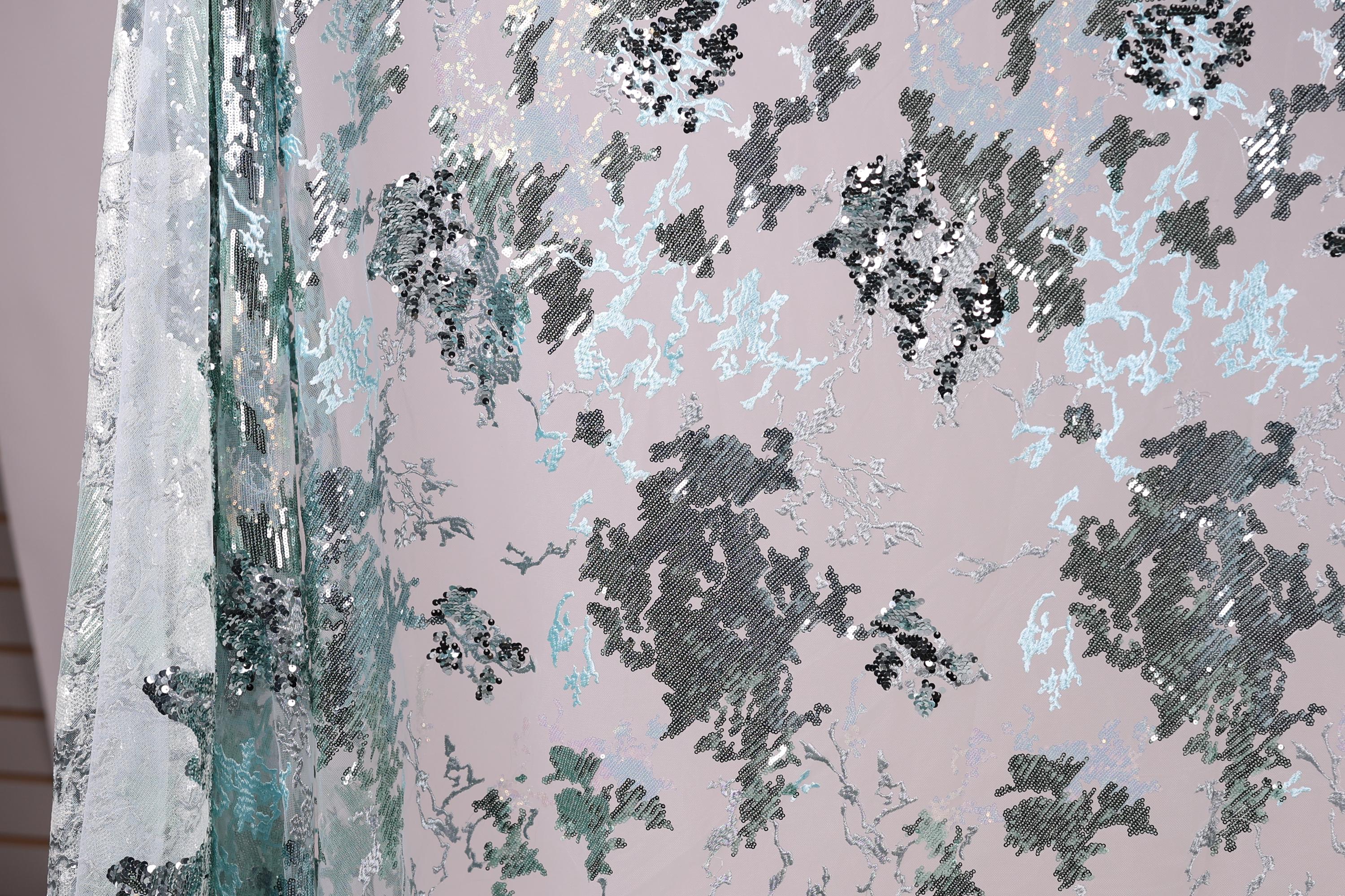 a close-up view of a fabric with a floral pattern in shades of blue and gray. The fabric appears to be made of a shiny, metallic material, giving it a textured and visually interesting appearance.