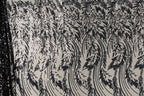 a close-up view of a fabric with a repeating pattern of black and white floral designs. The pattern consists of various shapes and lines, creating a visually striking and intricate design. The fabric appears to be made of a material with a metallic or sequined texture, adding a touch of sparkle and elegance to the overall appearance.