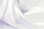 The image is a close-up of a soft, flowing white fabric, possibly a satin or chiffon material. The fabric appears to be wrinkled, giving it a delicate and elegant appearance.
