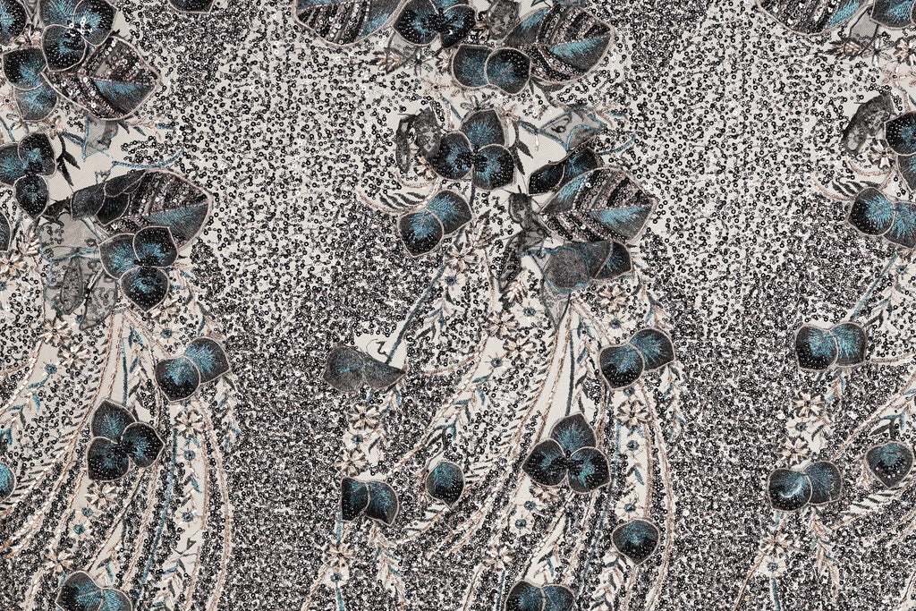 The image is a close-up of a fabric with a repeating pattern of blue and gray floral designs. The pattern consists of various shades of blue and gray flowers and leaves, creating a visually striking and intricate design. The fabric appears to be a textile or material with a textured surface, adding depth and dimension to the overall appearance.