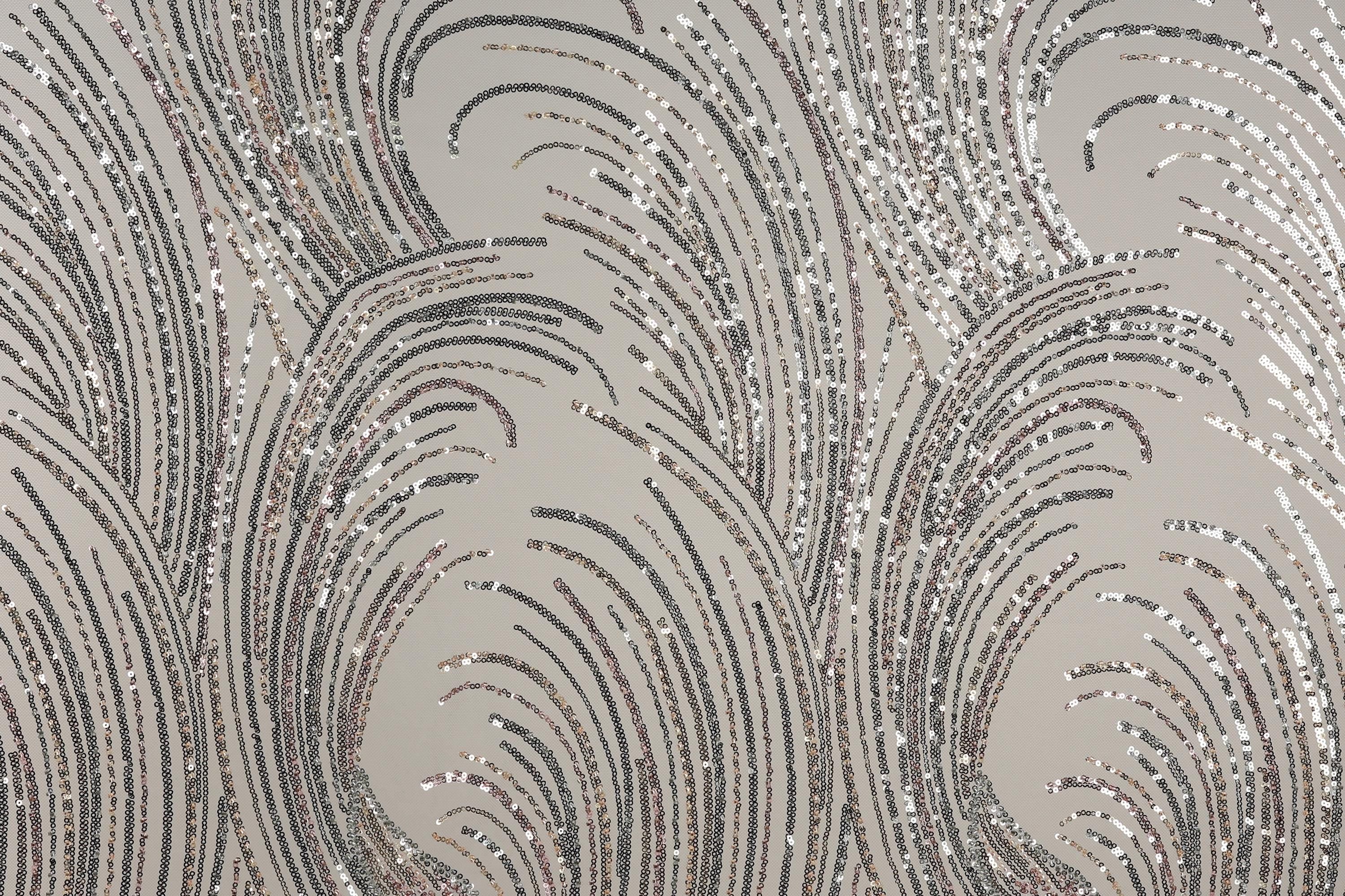 a close-up view of a fabric with a repeating pattern of silver and black sequin designs. The pattern consists of curved lines and swirls, creating a visually striking and intricate design. The fabric appears to be a light-colored material, possibly a type of lace or a similar decorative textile.