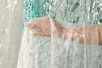 A hand is holding a piece of white lace with silver sequins, against a background of a green and white sequined curtain.