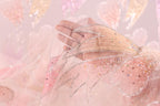 A hand is holding a delicate, sheer fabric adorned with sparkling rhinestones against a soft pink background.