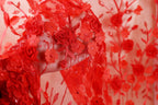 a close-up view of a hand holding a piece of red fabric adorned with delicate red flowers and leaves. The fabric appears to be a sheer, lace-like material, with the flowers and leaves intricately woven into the fabric, creating a visually striking and elegant design.