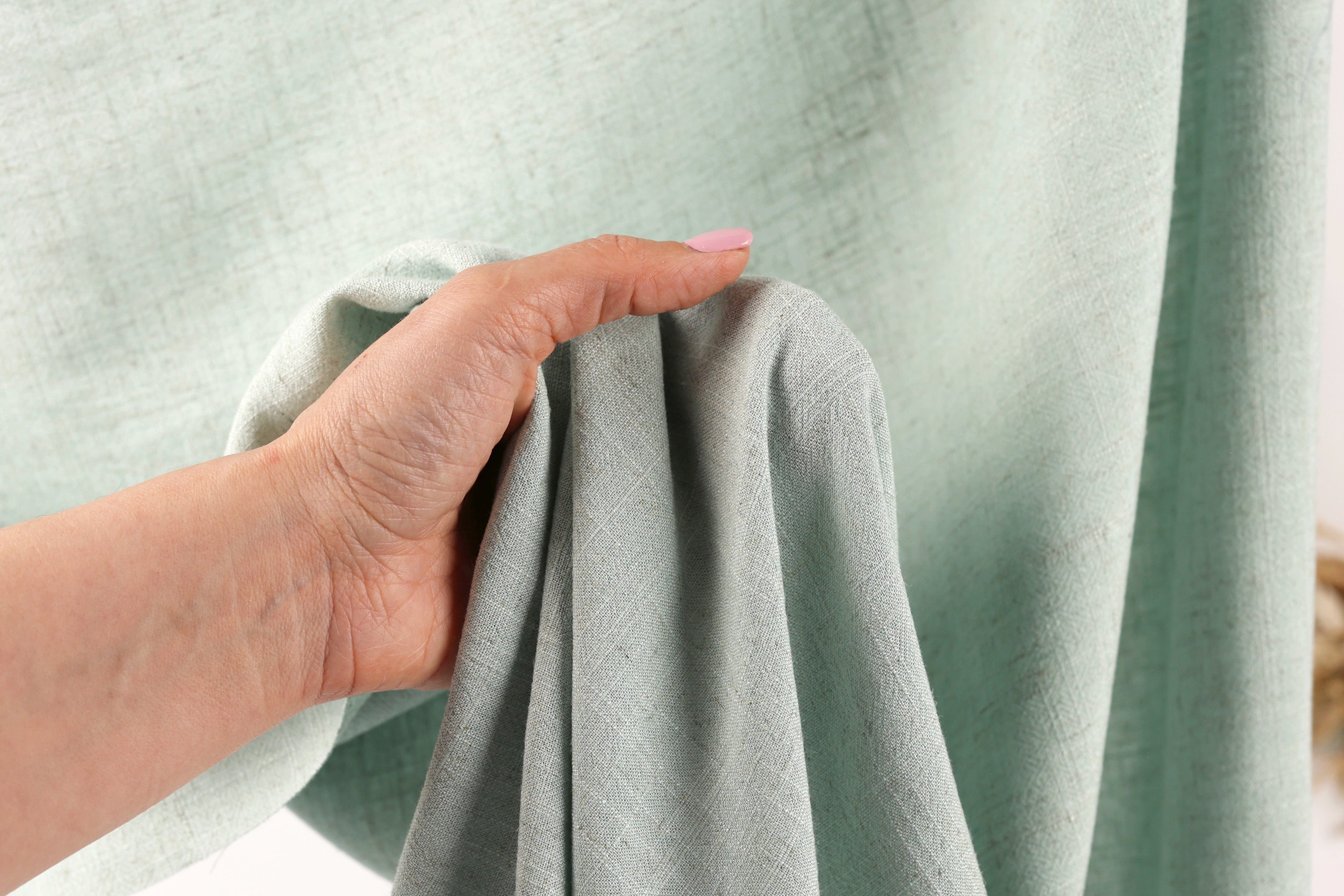 A hand is holding a light green fabric, possibly a curtain or textile, in front of a light green background.
