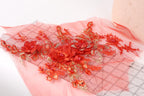 a close-up view of a delicate, intricate floral embroidery on a pink fabric. The embroidery is composed of numerous small red flowers, each adorned with a gold leaf, creating a visually striking and detailed design. The flowers are arranged in a way that they overlap and interlock, showcasing the skill and craftsmanship involved in the creation of this piece of textile art.
