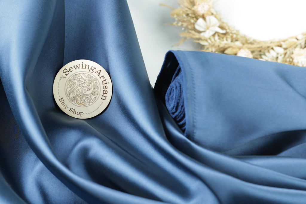 A round badge with the text "SewingArtisan Easy Shop" is placed on a blue fabric, with a wreath of dried flowers visible in the background.