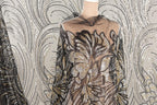 a mannequin wearing a dress with a sheer, sequined bodice and a floral pattern. The dress has a black and gold color scheme, with the bodice being black and the skirt being gold. The mannequin is positioned in front of a wall with a patterned design, creating a visually appealing contrast between the mannequin and the background.