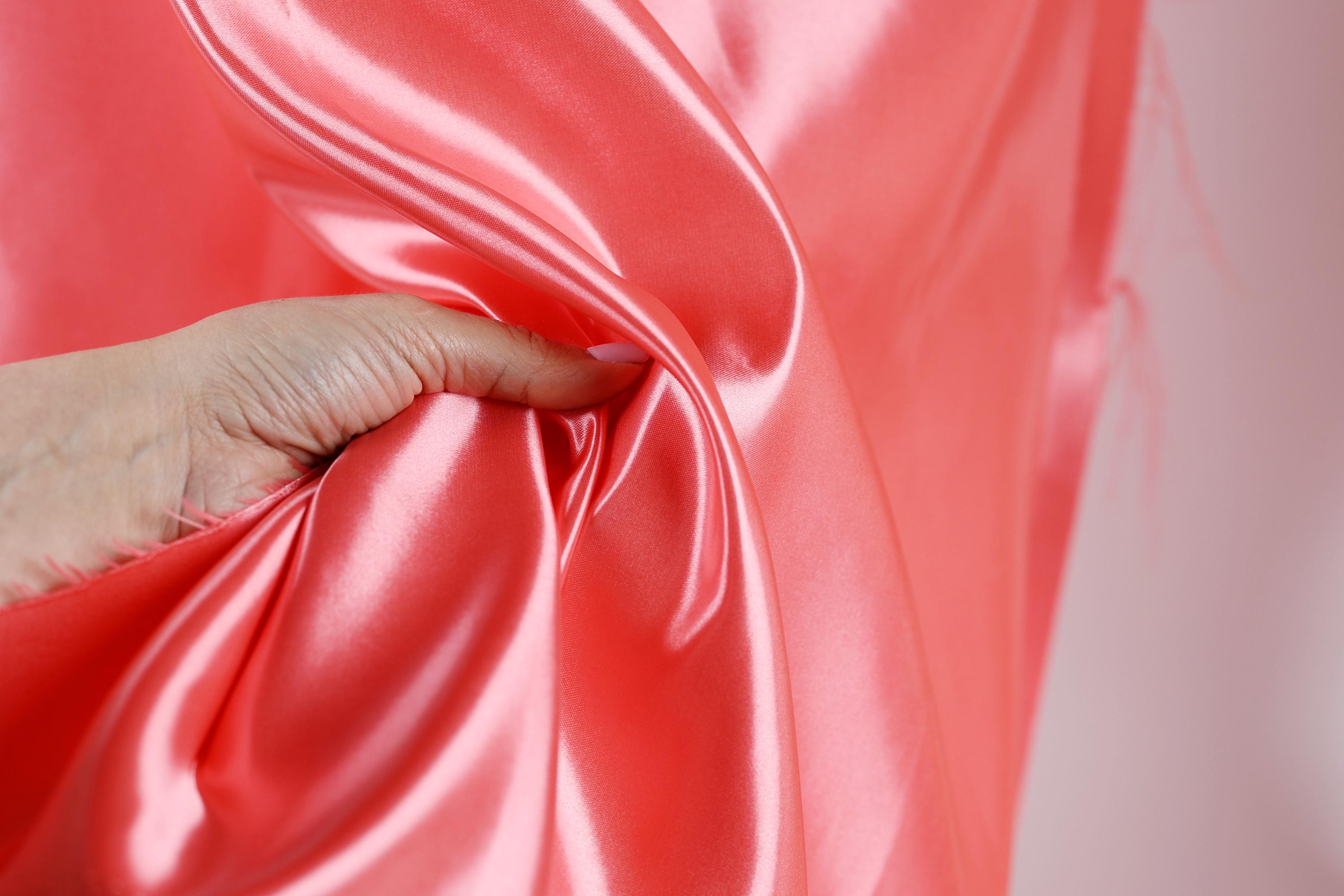 A hand is holding a piece of bright pink satin fabric, with the fabric being crumpled and wrinkled.