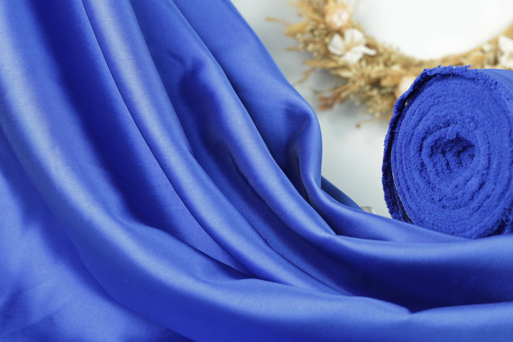 a close-up view of a blue fabric, possibly silk, with a rolled-up piece of the fabric visible in the background.