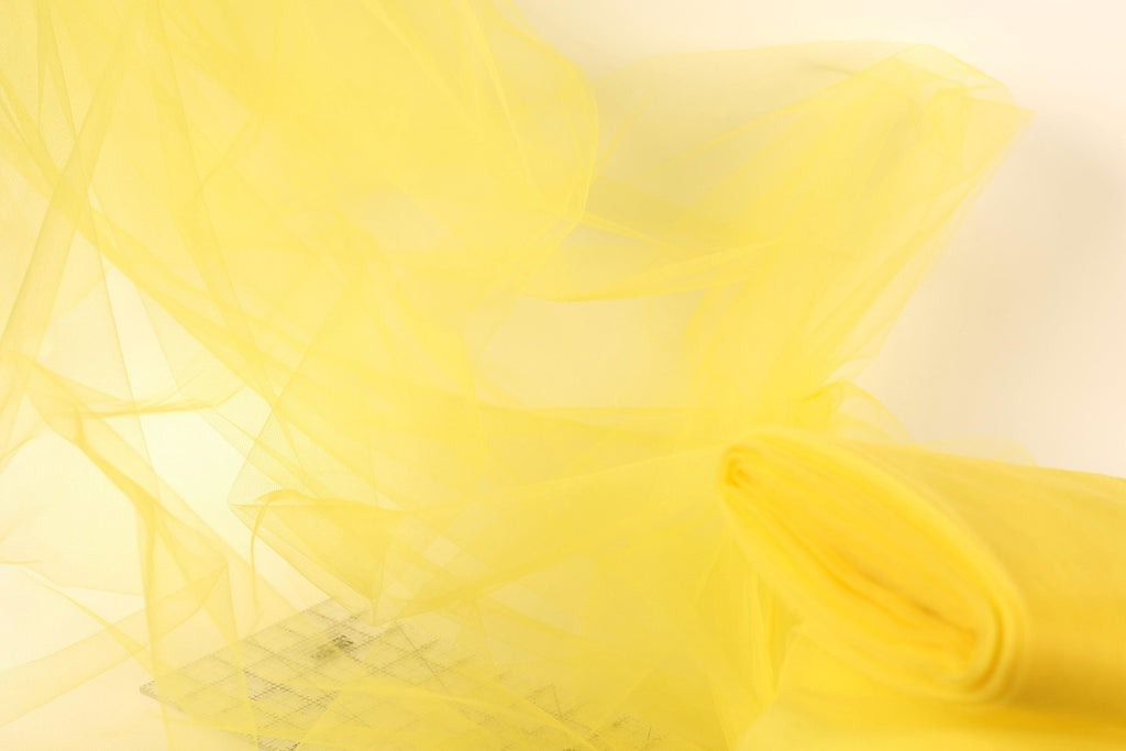 The image is a close-up of a yellow fabric, possibly tulle, with a soft, flowing texture. The fabric appears to be made of a sheer material, giving it a delicate and airy appearance. The background is a plain white color, which contrasts with the vibrant yellow of the fabric and draws attention to the fabric itself.