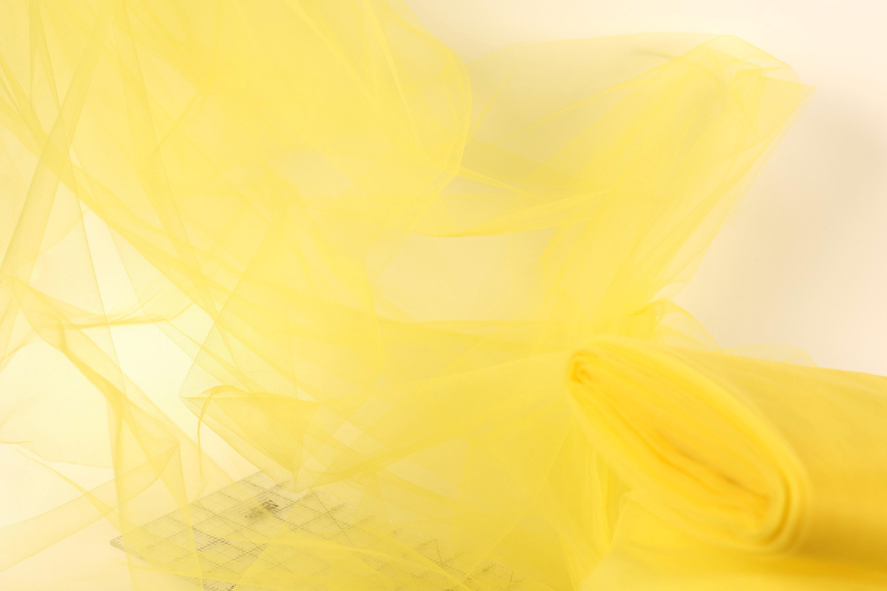 The image is a close-up of a yellow fabric, possibly tulle, with a soft, flowing texture. The fabric appears to be made of a sheer material, giving it a delicate and airy appearance. The background is a plain white color, which contrasts with the vibrant yellow of the fabric and draws attention to the fabric itself.
