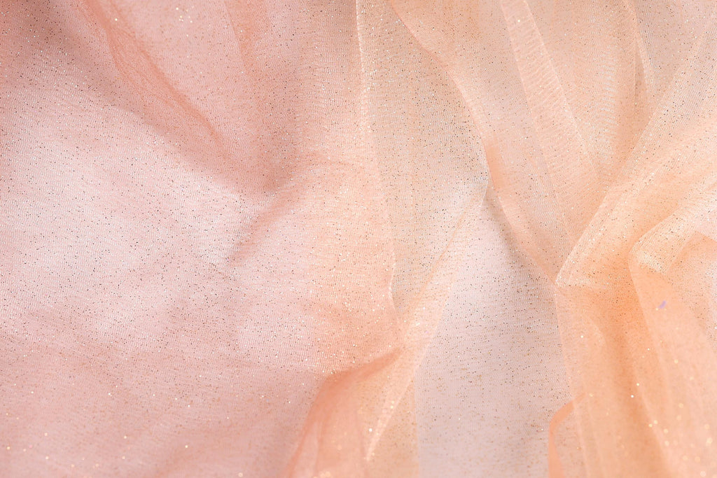 The image is a close-up of a fabric with a light peach or peach-colored texture, adorned with small, glittery particles or specks that create a sparkling effect.