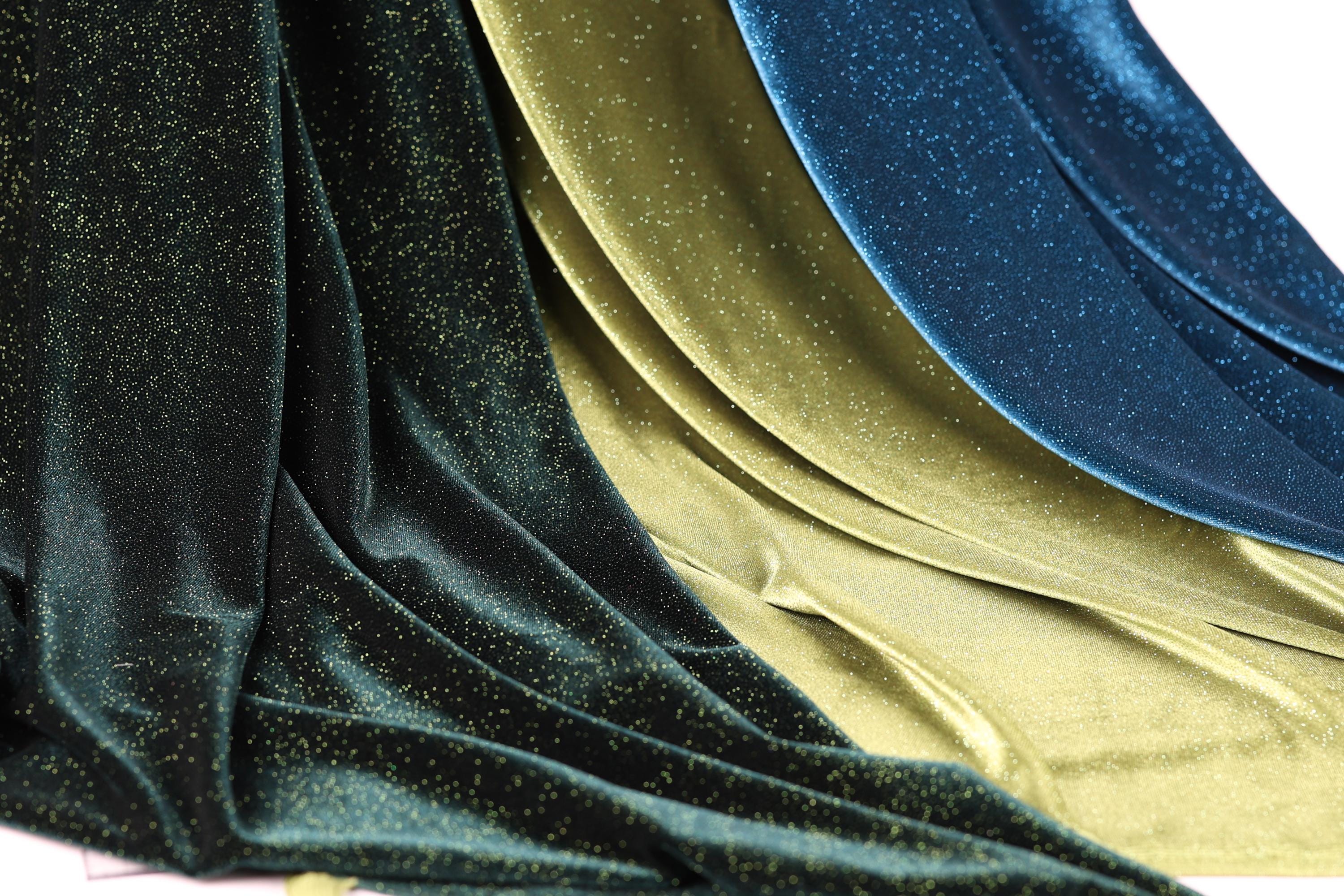 a close-up view of a fabric with a shimmering gold and blue glittery texture. The fabric appears to be made of a shiny material, possibly silk or velvet, and has a flowing, draped appearance. The colors of the fabric are gold and blue, creating a visually striking contrast.