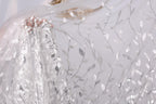 a close-up view of a delicate, sheer fabric with a floral pattern. The fabric appears to be made of sheer lace or chiffon, with a delicate, intricate design of leaves and flowers. The fabric is being held by a hand, and the background is a plain white color, which allows the fabric to stand out prominently.