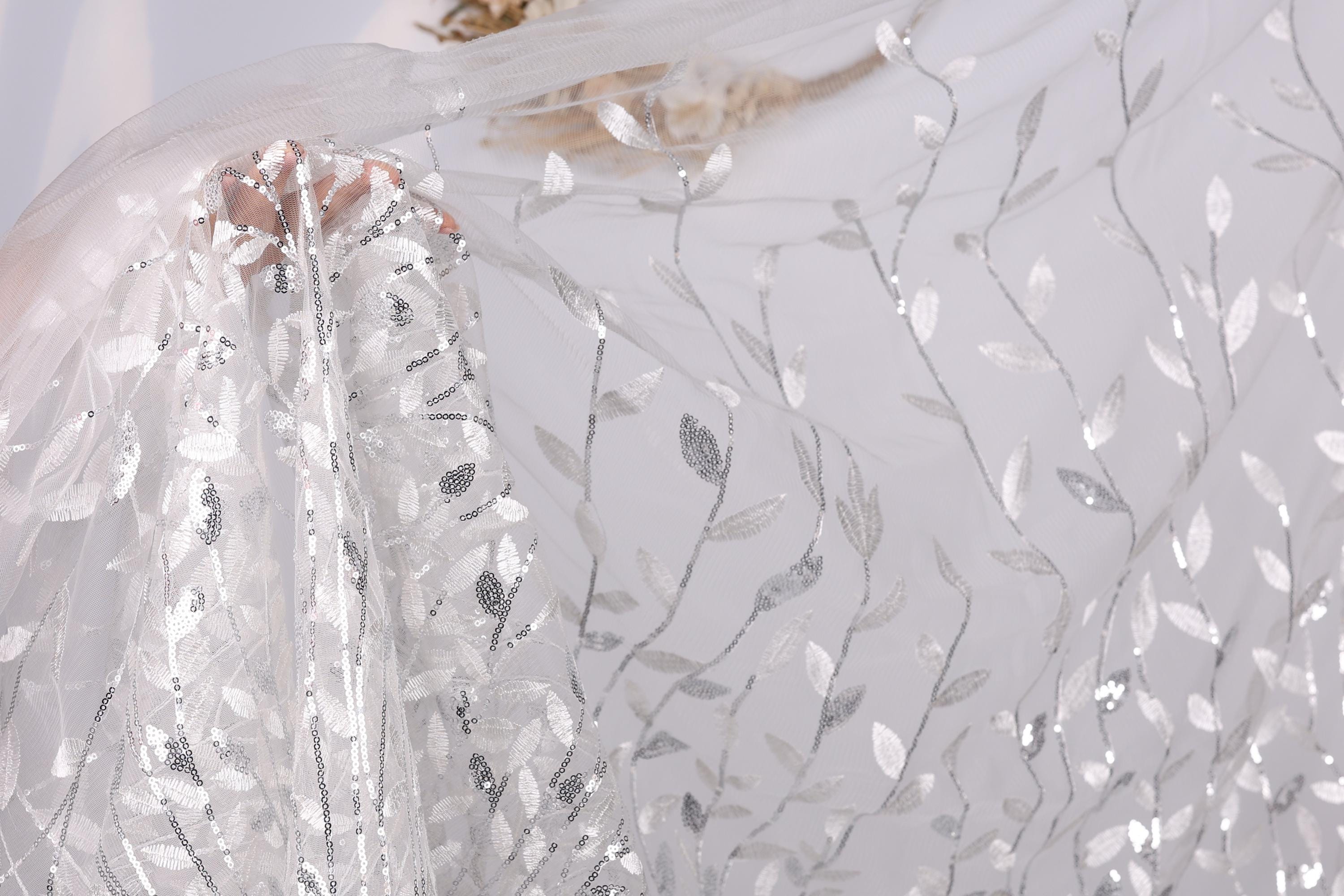 a close-up view of a delicate, sheer fabric with a floral pattern. The fabric appears to be made of sheer lace or chiffon, with a delicate, intricate design of leaves and flowers. The fabric is being held by a hand, and the background is a plain white color, which allows the fabric to stand out prominently.