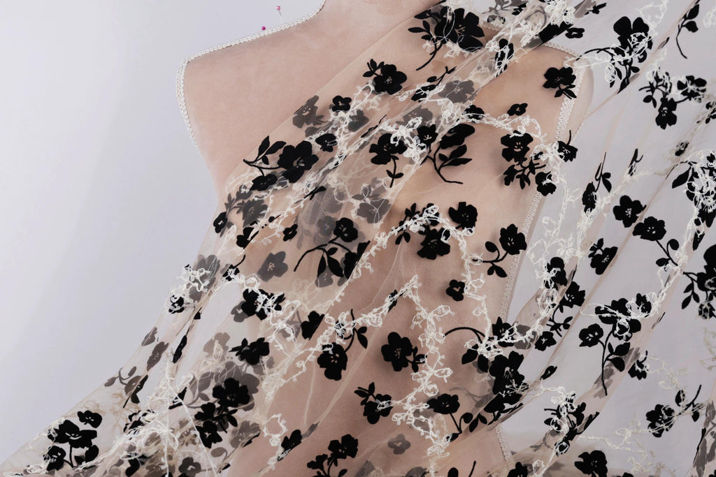 a close-up view of a delicate, sheer fabric with a black floral pattern. The fabric appears to be a sheer lace or chiffon material, with a floral design that includes black flowers and leaves. The fabric is displayed against a plain white background, which highlights the intricate details and textures of the fabric.
