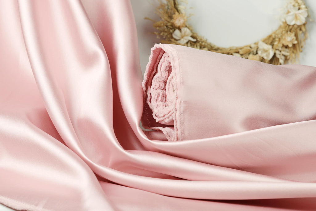A close-up view of a soft, pink satin fabric, possibly a sheer or satin material, with a floral wreath in the background.