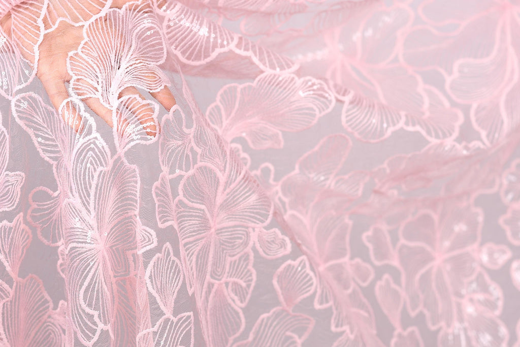 A close-up view of a delicate, pink lace fabric with intricate, repeating floral patterns.