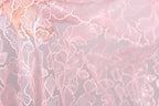 A close-up view of a delicate, pink lace fabric with intricate, repeating floral patterns.