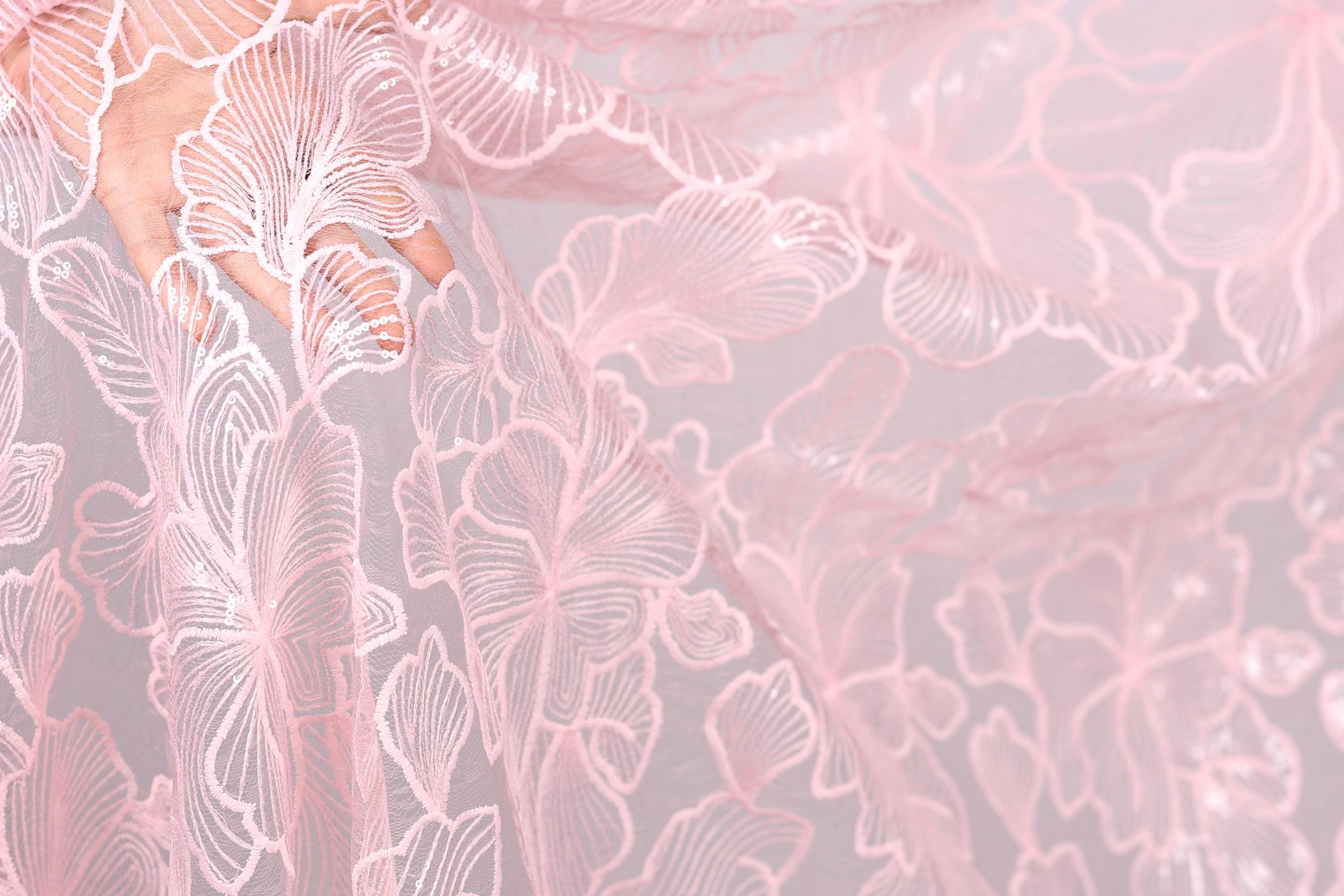 A close-up view of a delicate, pink lace fabric with intricate, repeating floral patterns.