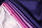 a close-up view of a fabric with a gradient of colors, transitioning from a deep purple to a lighter shade of purple. The fabric appears to be made of satin or a similar smooth, shiny material.