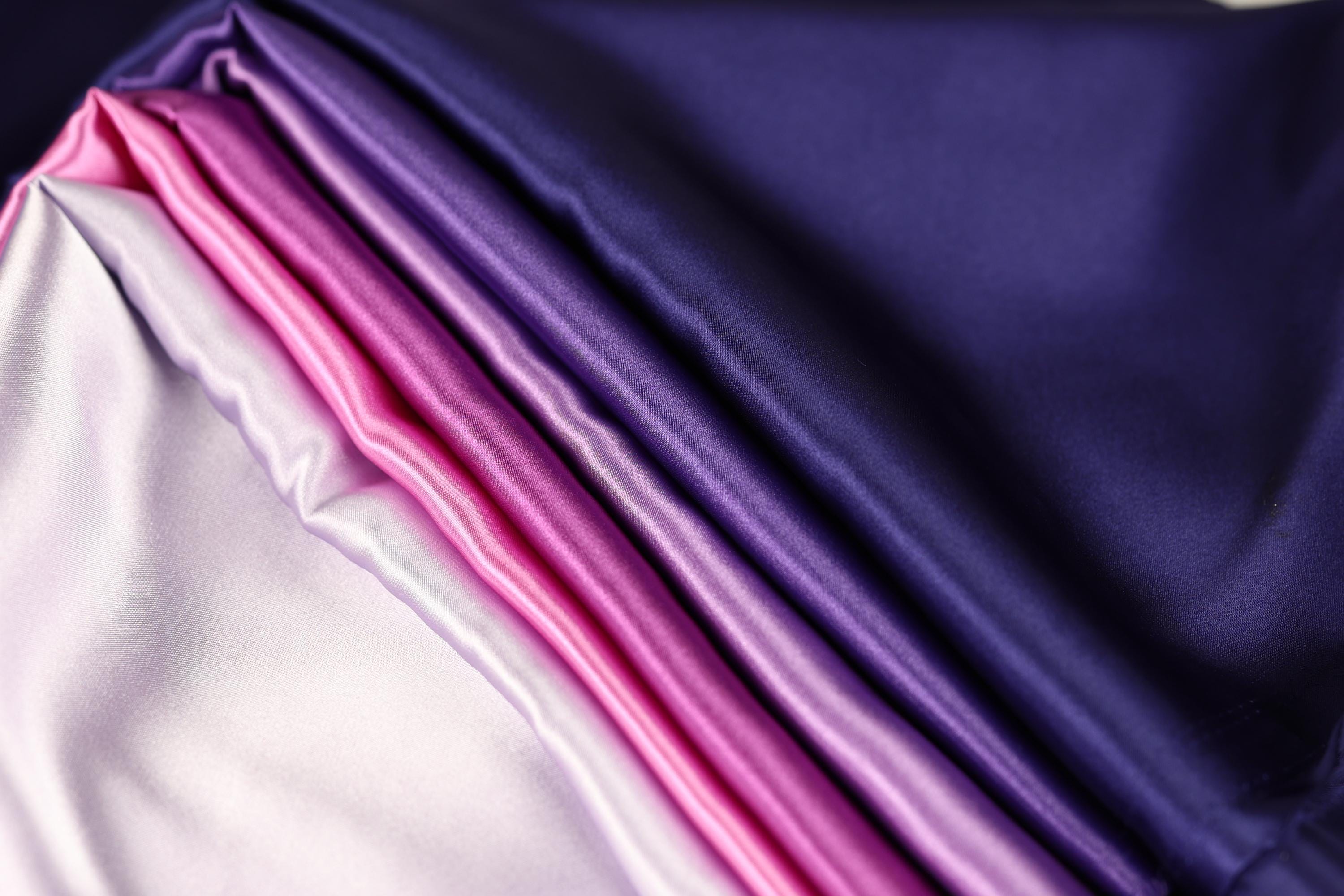 a close-up view of a fabric with a gradient of colors, transitioning from a deep purple to a lighter shade of purple. The fabric appears to be made of satin or a similar smooth, shiny material.