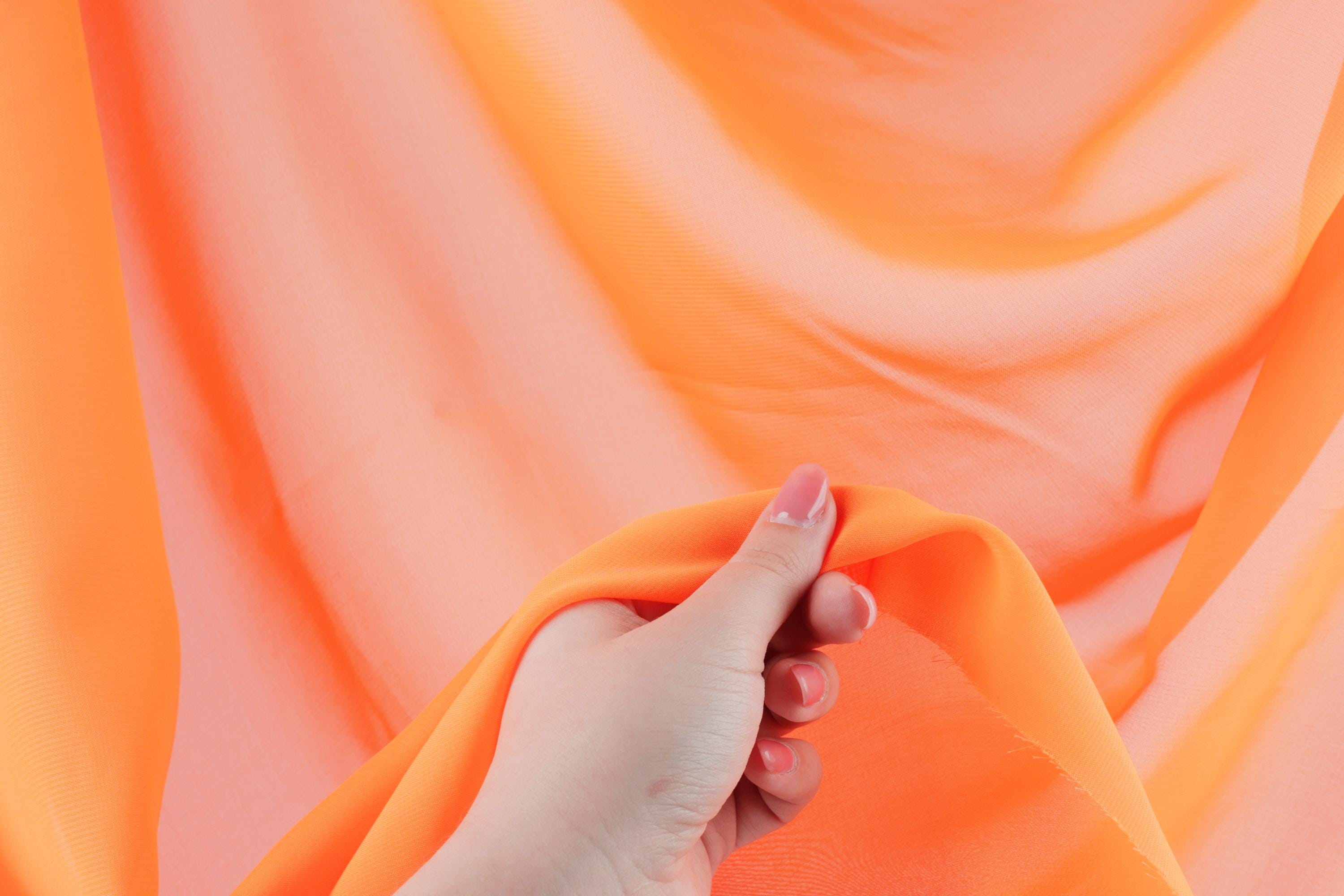 A hand is holding a piece of orange fabric, which appears to be a soft, flowing material.